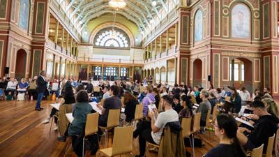 A group of budding artists sis on chairs under the dome of the Royal Exhibition Building for a drawing class.