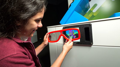 A person holds a large pair of red 3D glasses up to a small screen built into an exhibit. The screen displays a colourful, cartoon‑style image. The person stands beside a white and blue interactive display unit. 
