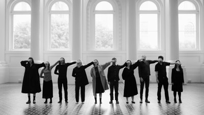 Line of performers covering one another’s eyes in a spacious hall with arched windows