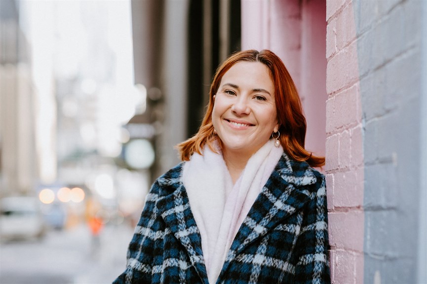 Zoë Condliffe with red hair leaning against a colourful brick wall in a winter jacket 