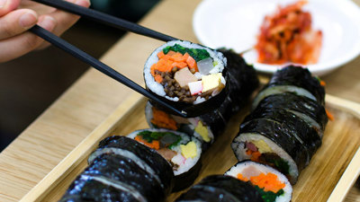  A close up of a hand holding chopsticks and picking up a piece of rice rolled in seaweed, with meat and vegetables inside. In the background is a plate of red pickled vegetables