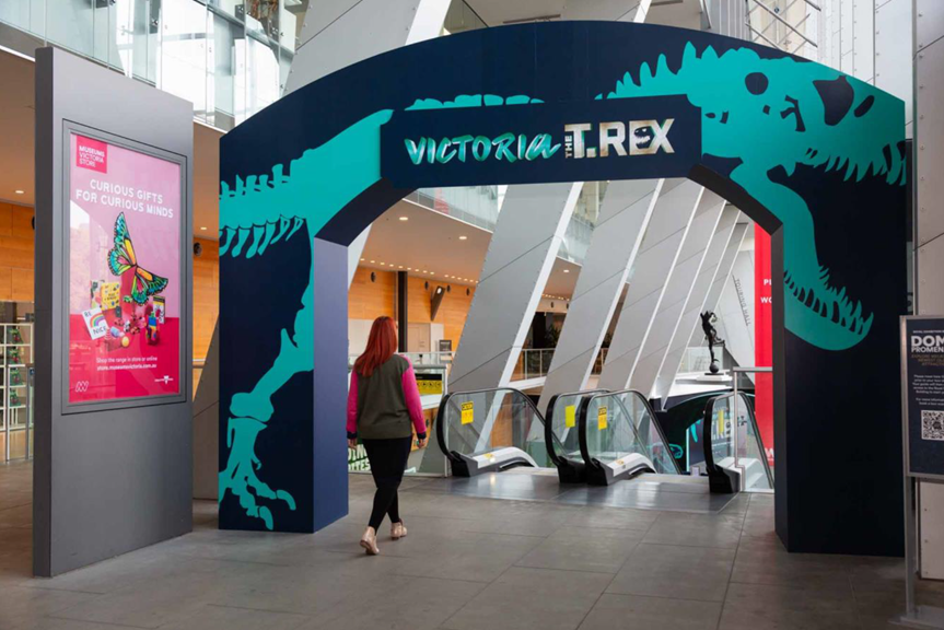 A person about to walk through the entrance and down the escalators leading to the Victoria the T. rex exhibition.