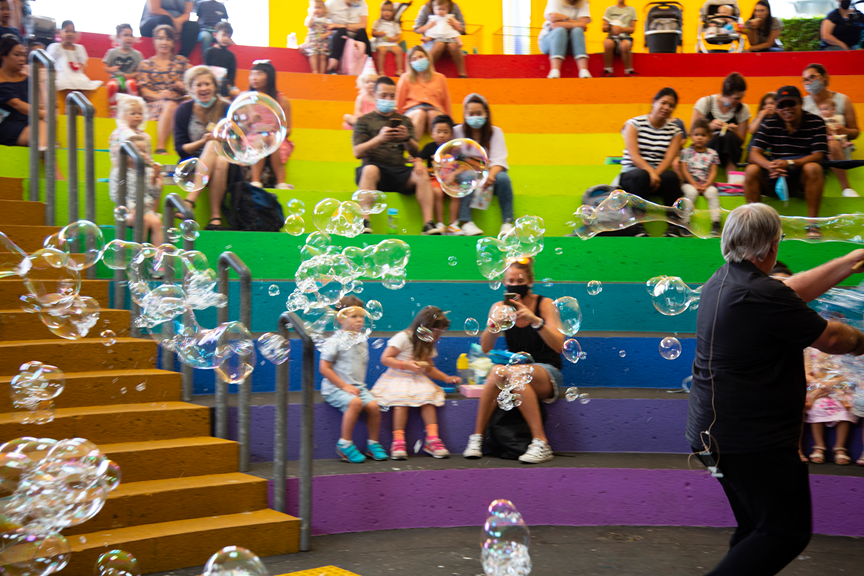 A performer dressed all in black makes many bubbles. They are trailing behind the presenter. Some are singles and others have clustered. A rainbow of tiered seating is scattered with families watching happily.